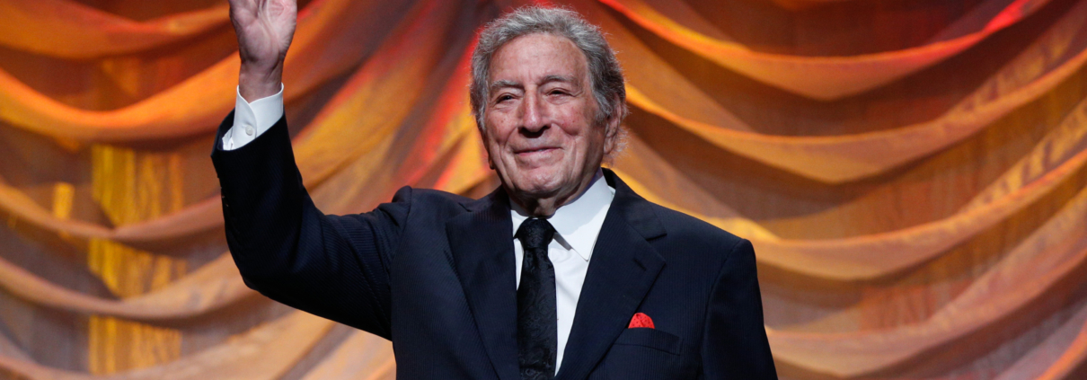 Singer Tony Bennett waves to a crowd while standing in front of an orange curtain.