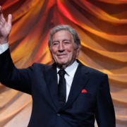 Singer Tony Bennett waves to a crowd while standing in front of an orange curtain.