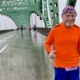 An elderly man in an orange shirt runs across a bridge on a rainy day.