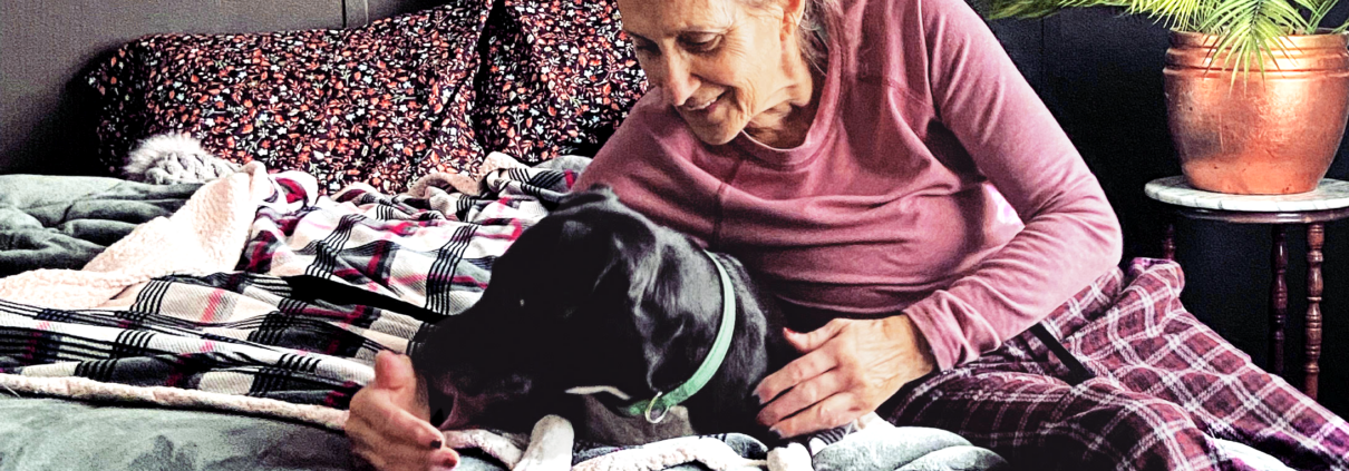 An older woman snuggles with a black dog on a bed.