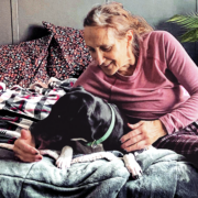 An older woman snuggles with a black dog on a bed.