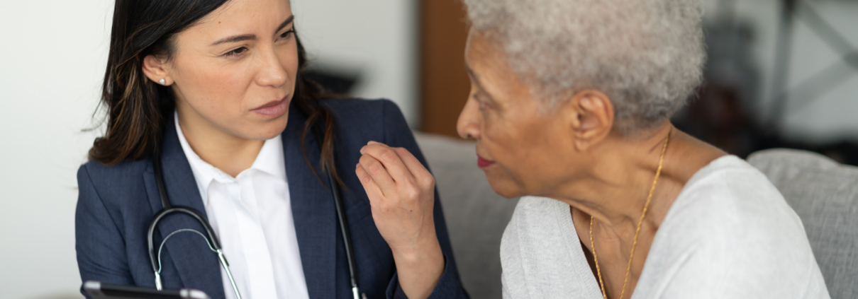 A young woman doctor in a white blouse and blue blazer has a stethoscope around her neck and holds a digital tablet. She is sitting and facing an older Black woman as they talk.