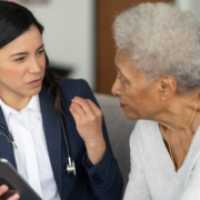 A young woman doctor in a white blouse and blue blazer has a stethoscope around her neck and holds a digital tablet. She is sitting and facing an older Black woman as they talk.