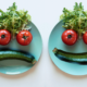 Two turquoise dinner plates sit side by side on a plain background. A cucumber, two tomatoes and lettuce on each plate are arranged to make a face. One face is smiling; the other is frowning.