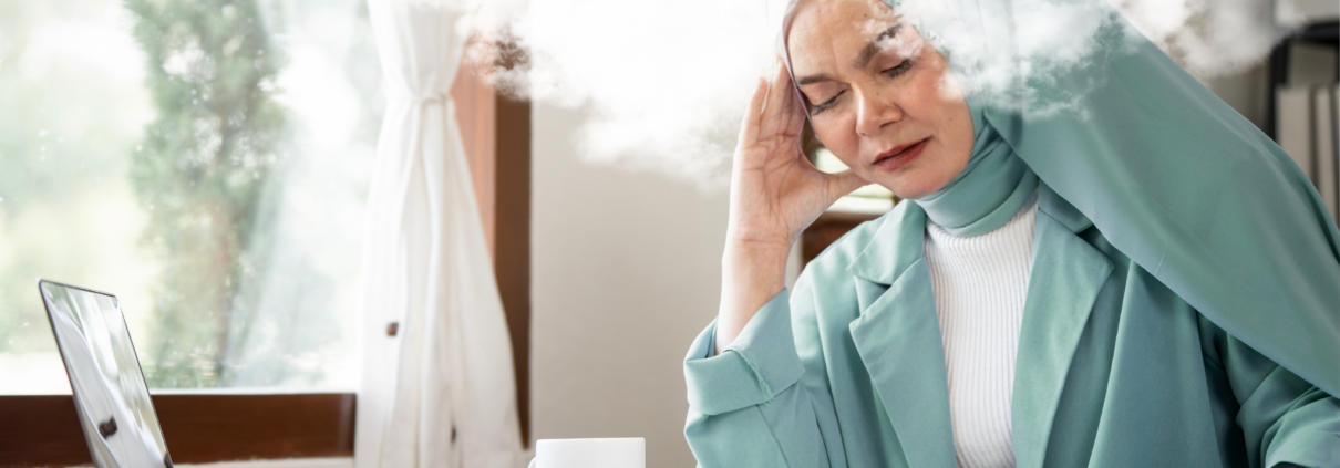 A woman in a mint green hijab, white ribbed shirt and mint green blazer closes her eyes and leans her head on her right hand on her desk, which has a few papers, coffee mug and laptop. An cloud of steam appears to surround her head, illustrating the concept of brain fog.