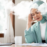 A woman in a mint green hijab, white ribbed shirt and mint green blazer closes her eyes and leans her head on her right hand on her desk, which has a few papers, coffee mug and laptop. An cloud of steam appears to surround her head, illustrating the concept of brain fog.