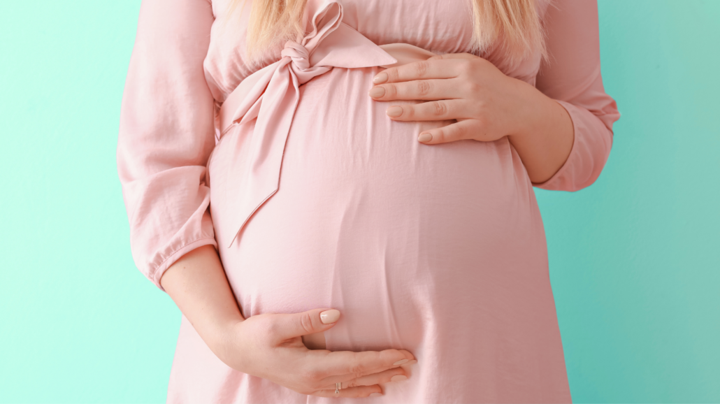 A white pregnant woman, shown only from the chest down to her belly, is wearing a pale pink dress and facing the camera while holding her hands over her belly.