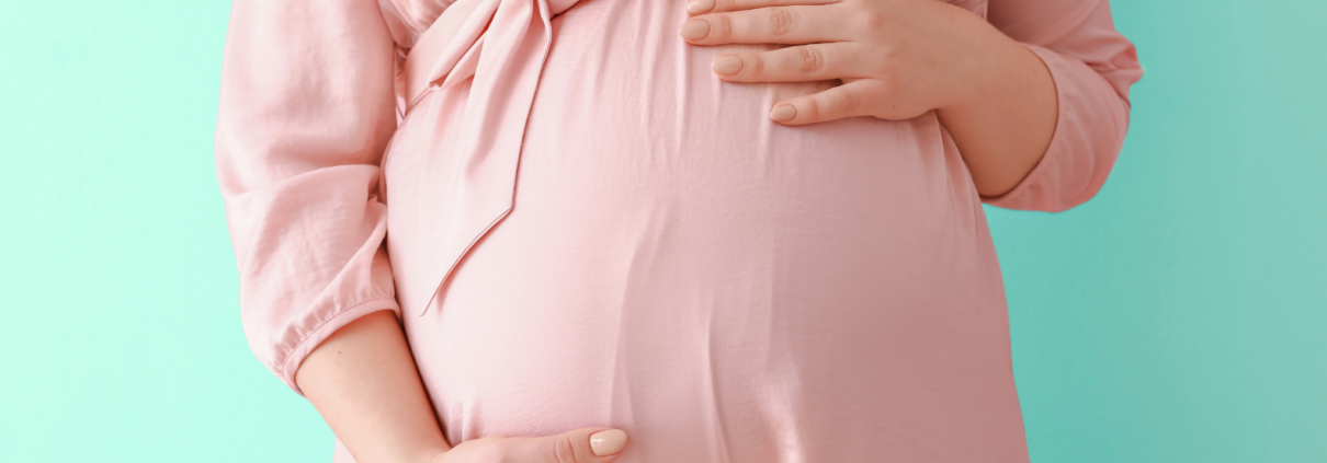 A white pregnant woman, shown only from the chest down to her belly, is wearing a pale pink dress and facing the camera while holding her hands over her belly.