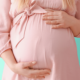 A white pregnant woman, shown only from the chest down to her belly, is wearing a pale pink dress and facing the camera while holding her hands over her belly.