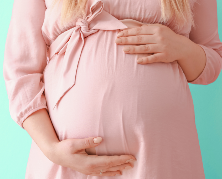 A white pregnant woman, shown only from the chest down to her belly, is wearing a pale pink dress and facing the camera while holding her hands over her belly.
