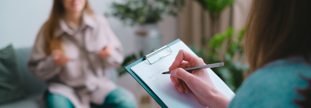 A white woman, who we only see from the back, holds a pen in her right hand while writing on a clipboard. Across from her, sitting on a couch is a white woman, who is out of focus and gesturing with her hands.