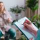 A white woman, who we only see from the back, holds a pen in her right hand while writing on a clipboard. Across from her, sitting on a couch is a white woman, who is out of focus and gesturing with her hands.