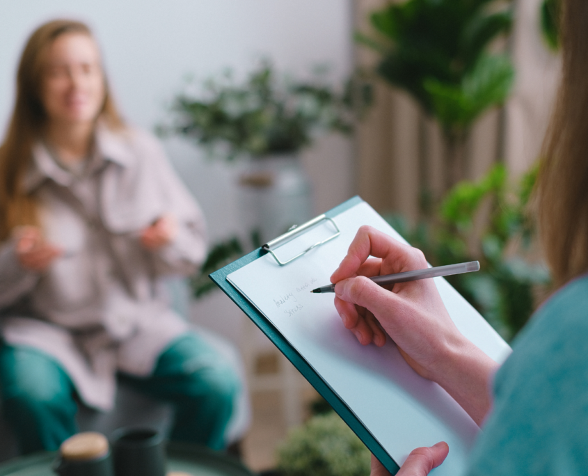 A white woman, who we only see from the back, holds a pen in her right hand while writing on a clipboard. Across from her, sitting on a couch is a white woman, who is out of focus and gesturing with her hands.