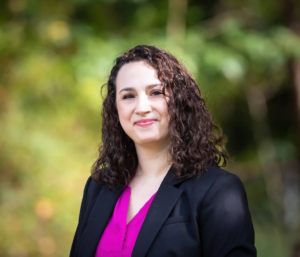 A professional portrait photo of Gillian Velmer. She is wearing a bright pink blouse with a black blazer. She is a white woman with dark brown curly hair hitting her shoulders and no bangs. She is smiling at the camera, and the background is a blurry green nature scene.