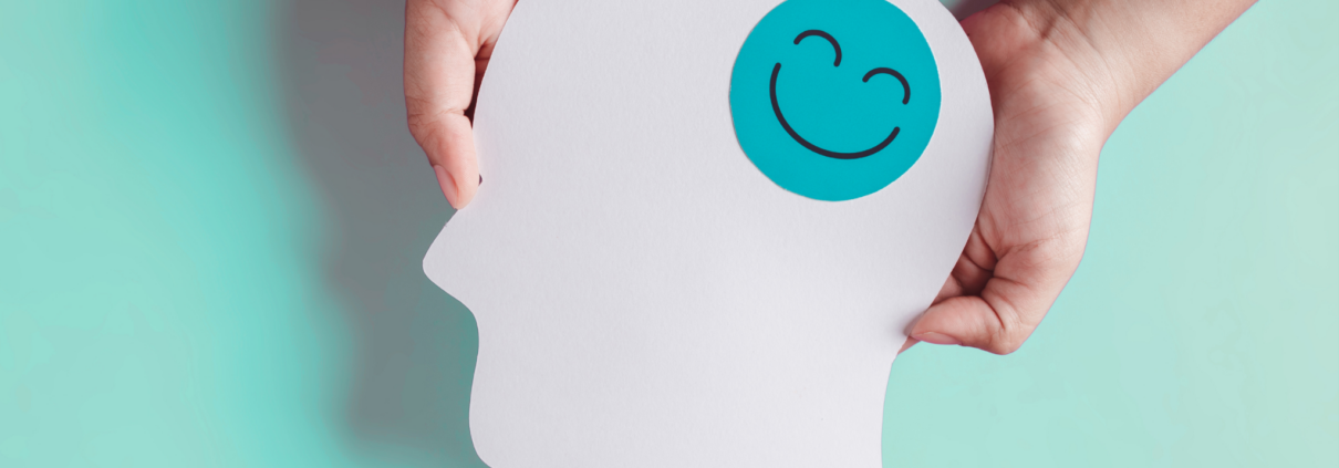 Two white hands hold a paper cutout of a human head, shown in profile, with a green happy face sticker on the head.
