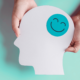 Two white hands hold a paper cutout of a human head, shown in profile, with a green happy face sticker on the head.
