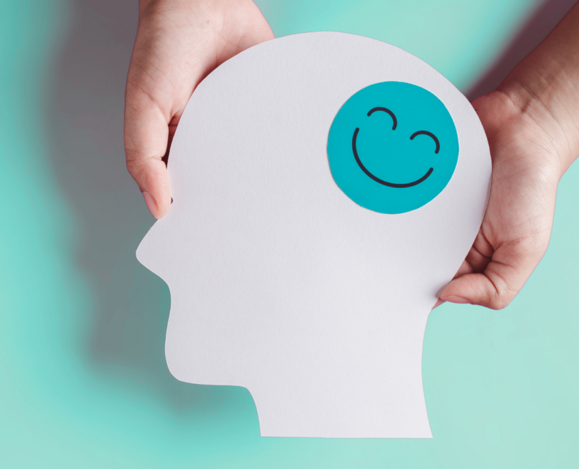 Two white hands hold a paper cutout of a human head, shown in profile, with a green happy face sticker on the head.