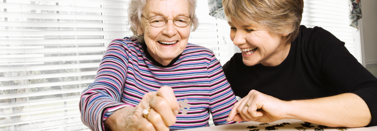 An elderly white woman and a middle-aged white woman smile and laugh together while working on a jigsaw puzzle.