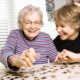 An elderly white woman and a middle-aged white woman smile and laugh together while working on a jigsaw puzzle.