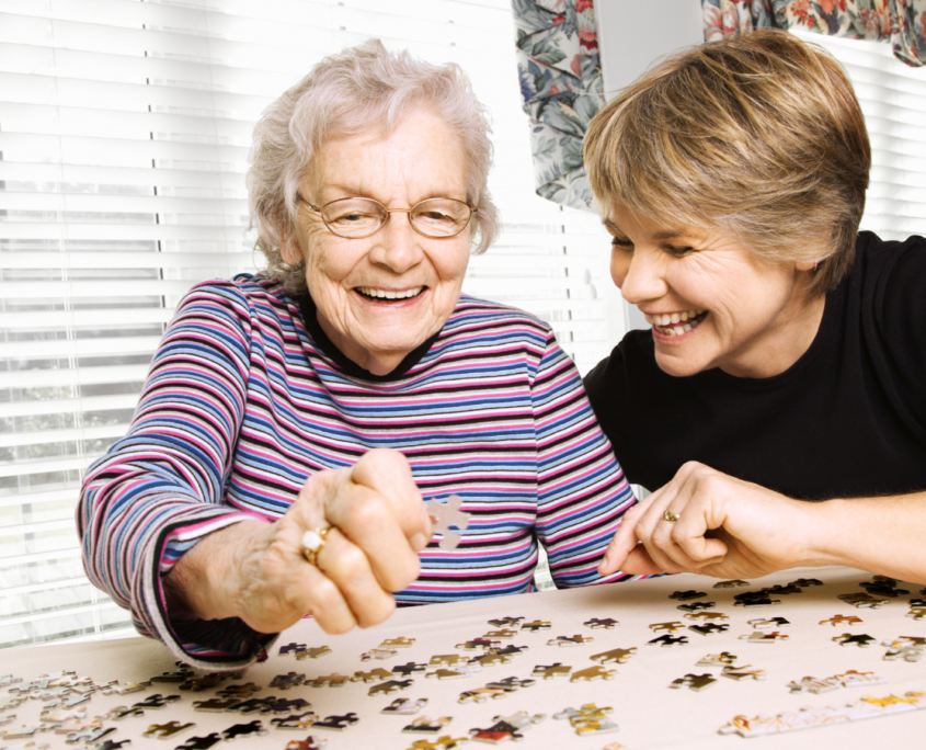 An elderly white woman and a middle-aged white woman smile and laugh together while working on a jigsaw puzzle.
