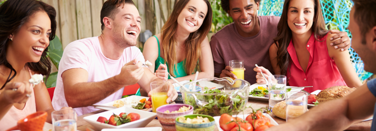A group of six ethnically diverse friends in their 20s and 30s sit around a table outdoors laughing and eating a healthy array of salads and fruits and vegetables.