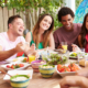 A group of six ethnically diverse friends in their 20s and 30s sit around a table outdoors laughing and eating a healthy array of salads and fruits and vegetables.
