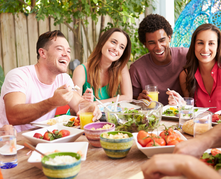 A group of six ethnically diverse friends in their 20s and 30s sit around a table outdoors laughing and eating a healthy array of salads and fruits and vegetables.