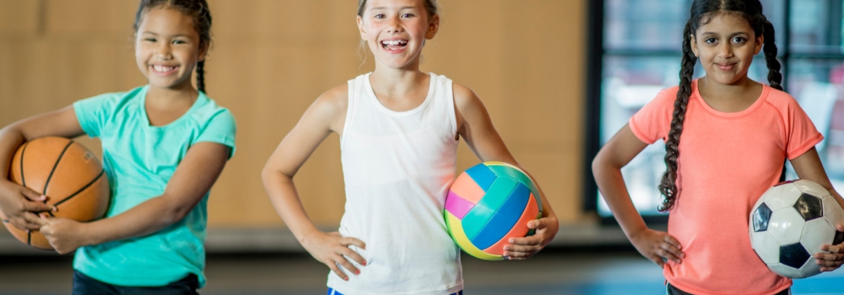 Three girls stand in a row, facing the camera and smiling. They are all wearing gyms shorts and T-shirts. One is holding a basketball, one a volleyball and one a soccer ball.