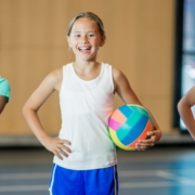 Three girls stand in a row, facing the camera and smiling. They are all wearing gyms shorts and T-shirts. One is holding a basketball, one a volleyball and one a soccer ball.