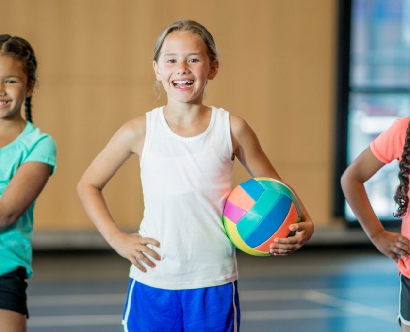Three girls stand in a row, facing the camera and smiling. They are all wearing gyms shorts and T-shirts. One is holding a basketball, one a volleyball and one a soccer ball.