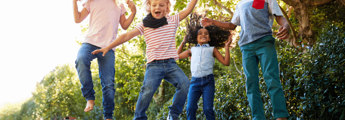 Four elementary-age kids jump up and down on a trampoline and laugh and smile at the camera.