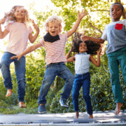 Four elementary-age kids jump up and down on a trampoline and laugh and smile at the camera.