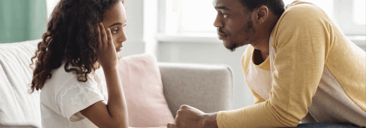 A Black girl about age 12 looks sad and holds her right arm to her forehead, while a Black man sitting across from her, facing her, leans forward and holds her hand with both his hands.