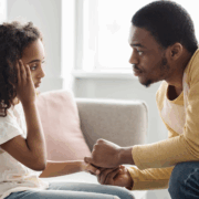 A Black girl about age 12 looks sad and holds her right arm to her forehead, while a Black man sitting across from her, facing her, leans forward and holds her hand with both his hands.