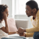 A Black girl about age 12 looks sad and holds her right arm to her forehead, while a Black man sitting across from her, facing her, leans forward and holds her hand with both his hands.