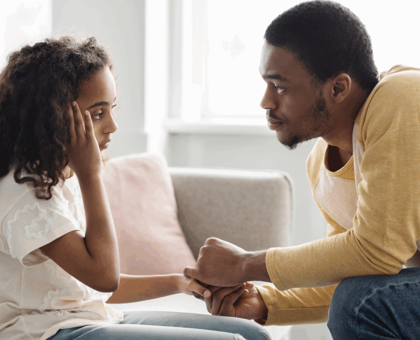 A Black girl about age 12 looks sad and holds her right arm to her forehead, while a Black man sitting across from her, facing her, leans forward and holds her hand with both his hands.