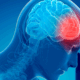 An illustration of a human head is shown in profile. Like an X-ray, we can see the spinal column and brain. Everything is in blue colors except for an area at the front of the brain that is in red. And the human his holding their hand to their head.