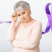 BW_Alz_women new An elderly white woman with short gray hour and wearing a light-colored peach top with long sleeves is looking down and holding her right hand to her head, as if in thought. A purple ribbon design is part of the background image.