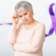 An elderly white woman with short gray hour and wearing a light-colored peach top with long sleeves is looking down and holding her right hand to her head, as if in thought. A purple ribbon design is part of the background image.
