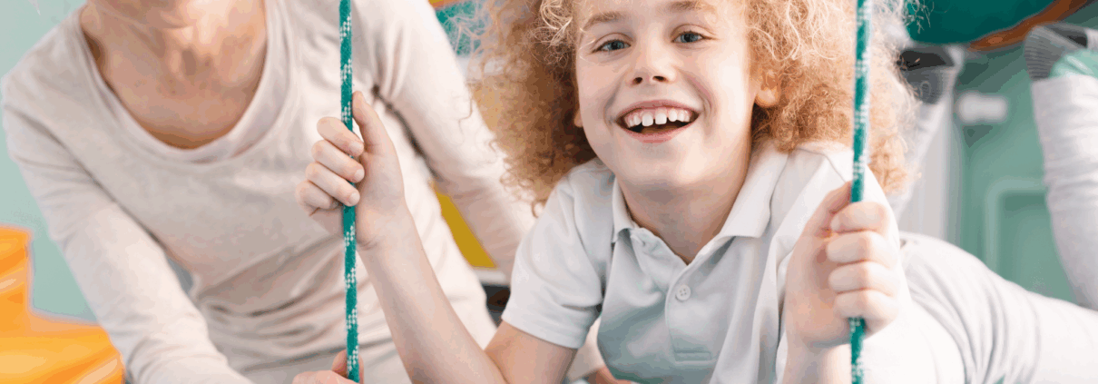 A child about age 7 with long frizzy reddish-blonde hair and wearing white pants and a white Polo shirt lays on their stomach on a swinging cushion-type mat while smiling at the camera. An adult blonde woman is looking at the child and smiling with her hand spotting them on the mat.