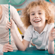 A child about age 7 with long frizzy reddish-blonde hair and wearing white pants and a white Polo shirt lays on their stomach on a swinging cushion-type mat while smiling at the camera. An adult blonde woman is looking at the child and smiling with her hand spotting them on the mat.