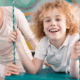 A child about age 7 with long frizzy reddish-blonde hair and wearing white pants and a white Polo shirt lays on their stomach on a swinging cushion-type mat while smiling at the camera. An adult blonde woman is looking at the child and smiling with her hand spotting them on the mat.
