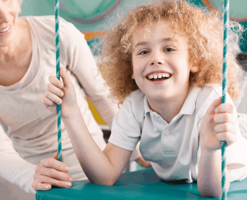 A child about age 7 with long frizzy reddish-blonde hair and wearing white pants and a white Polo shirt lays on their stomach on a swinging cushion-type mat while smiling at the camera. An adult blonde woman is looking at the child and smiling with her hand spotting them on the mat.