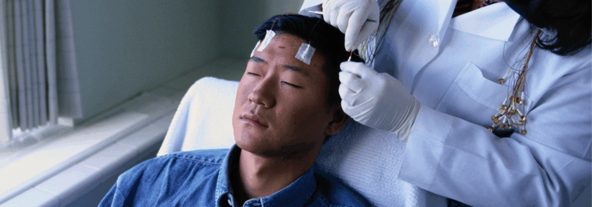A young Asian male lays semi-reclined on a hospital chair with his eyes closed while a technician in a white coat puts electrodes on his head.