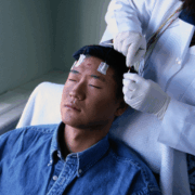 A young Asian male lays semi-reclined on a hospital chair with his eyes closed while a technician in a white coat puts electrodes on his head.