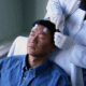 A young Asian male lays semi-reclined on a hospital chair with his eyes closed while a technician in a white coat puts electrodes on his head.