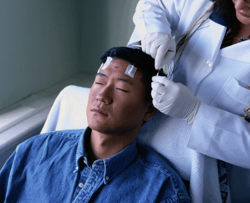 A young Asian male lays semi-reclined on a hospital chair with his eyes closed while a technician in a white coat puts electrodes on his head.