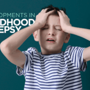 A young white boy around age 8 has his eyes closed and hands on the front of his head as if in pain or frustration. The image has the words "Developments in Childhood Epilepsy" on it.