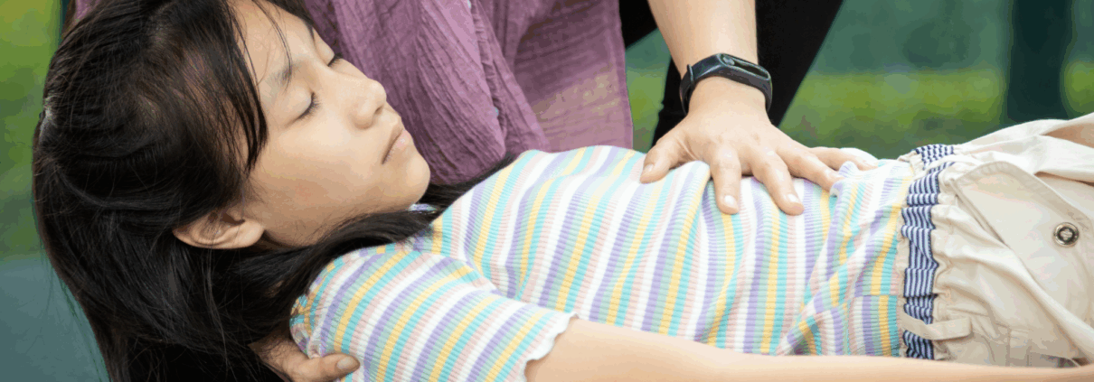 A girl about age 12 lies on the floor with her eyes closed. A woman puts her left hand on the girl's chest and cradles her head with her right hand.