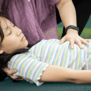 A girl about age 12 lies on the floor with her eyes closed. A woman puts her left hand on the girl's chest and cradles her head with her right hand.
